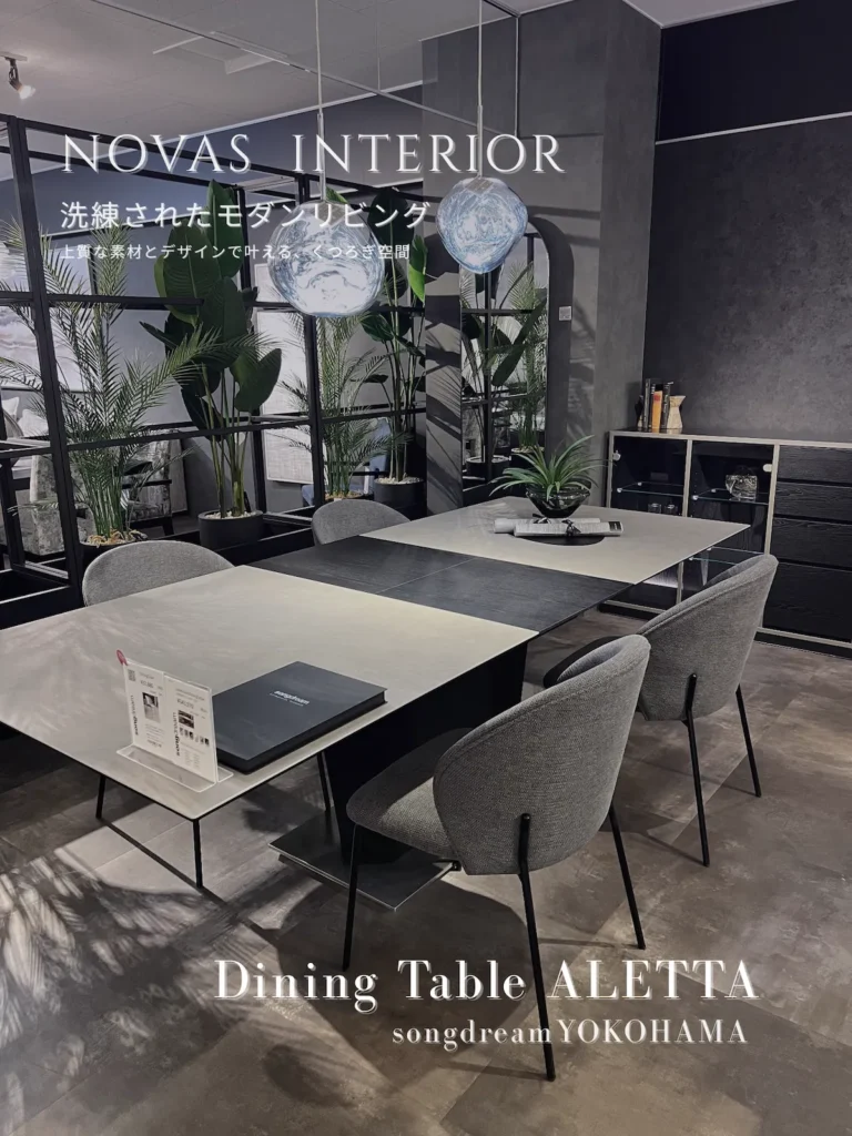 songdreamのショールーム展示。白と黒の天板を持つモダンなテーブルに、シンプルなグレーのチェアを合わせたダイニング。2つの青い球体ペンダントライトが吊るされ、背景には植物やガラスの仕切りがある。画像上部に「NOVAS INTERIOR...」の文字が重なる。