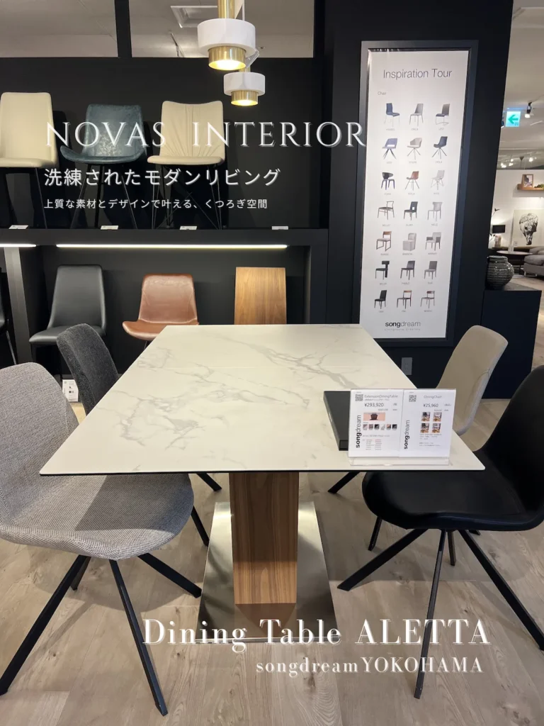 インテリアショップ「songdream」の展示。白の大理石模様と中央に木目が入った伸長式ダイニングテーブル「ALETTA」を中心に、グレー、木製、ベージュ、黒など異なるタイプのチェアが配置されている。背景には他のチェアの展示やプライスカードが見える。画像には「NOVAS INTERIOR Dining Table ALETTA 洗練されたモダンリビング...」の文字が重ねられている。